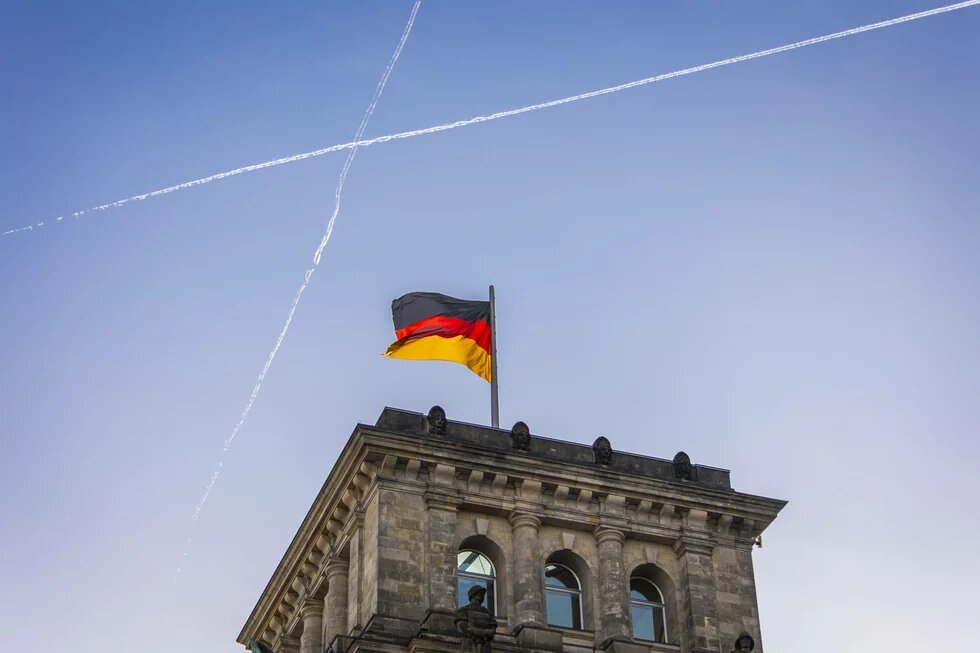 Ein Gebäudeturm des Reichstags in Berlin mit wehender deutscher Flagge. Am Himmel kreuzen sich zwei Kondensstreifen.